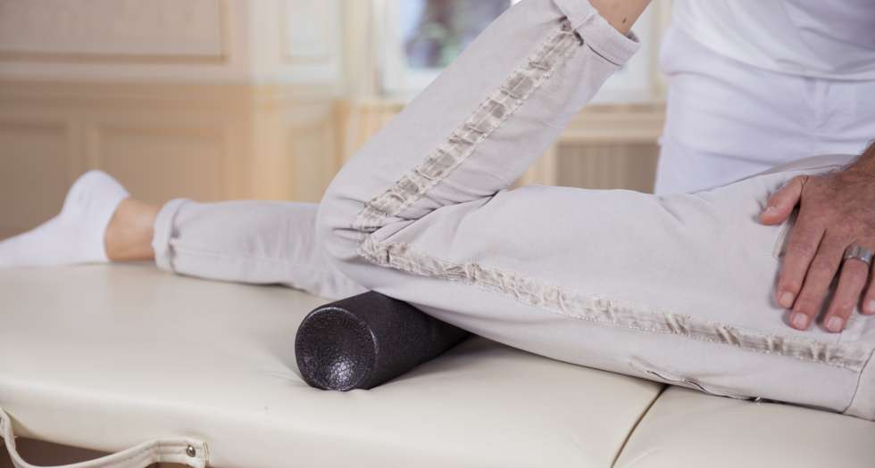 Faszienrollmassage Person liegt auf einer Liege und rollt ihren Oberschenkel auf einer Faszienrolle